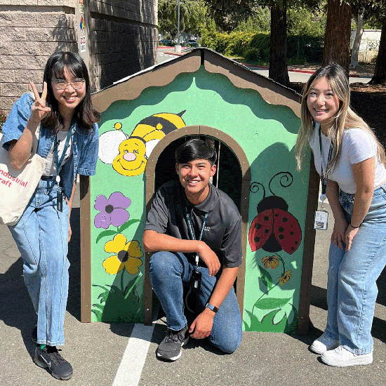 Two girls and a guy pose for the camera in front of a brightly-painted wooden playhouse