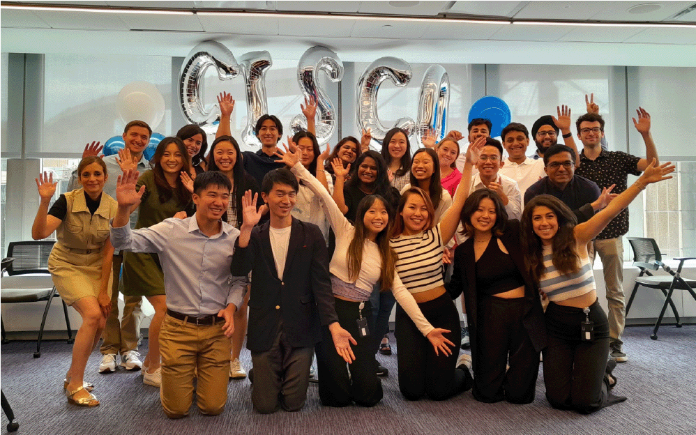 A large group of young people, the Cisco interns, pose in front of a bunch of balloons that read 