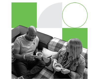 Man and woman, each holding a cup of coffee, are sitting on a couch, smiling at each other