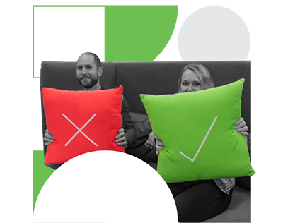A man, sitting on a couch, holds a pillow with an X printed on it. A woman sits next to him, holding a similar pillow with a checkmark printed on it