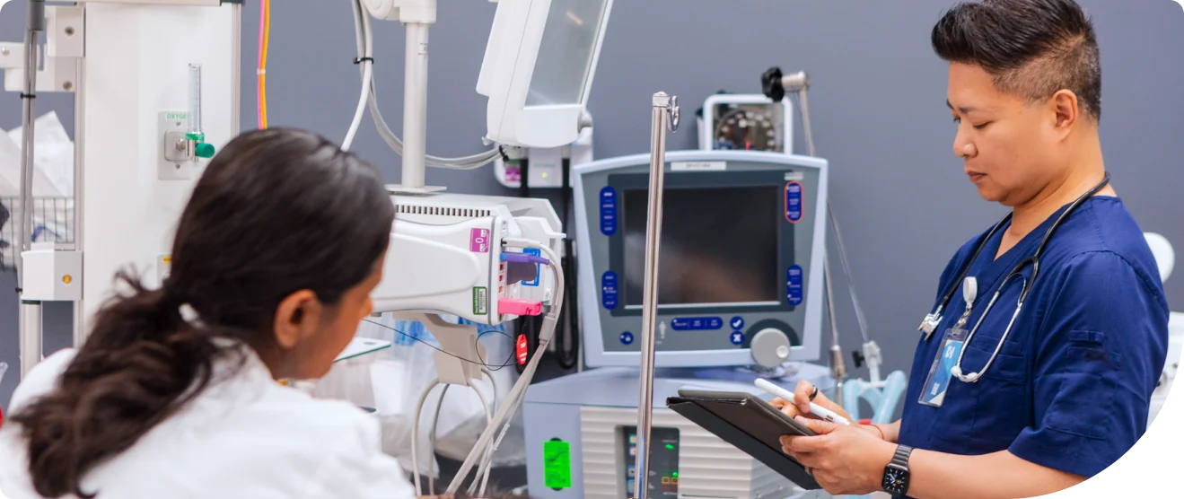 A healthcare worker in scrubs reviews information on a tablet beside medical monitors and equipment with a patient nearby