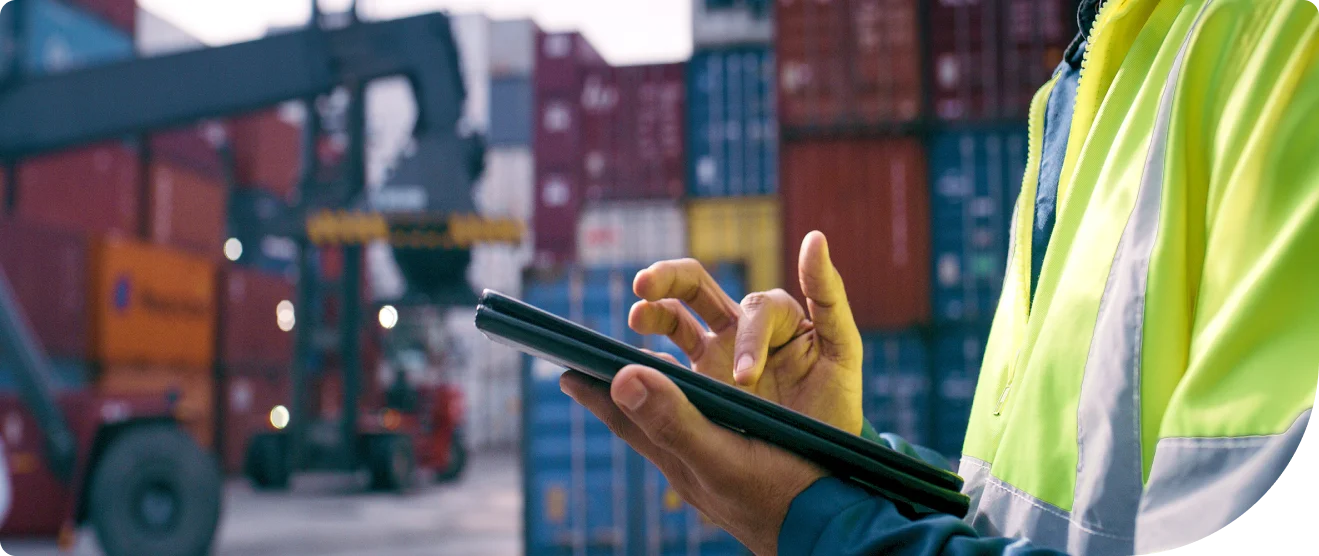 A worker in a high-visibility jacket uses a tablet in a shipping yard with stacked cargo containers