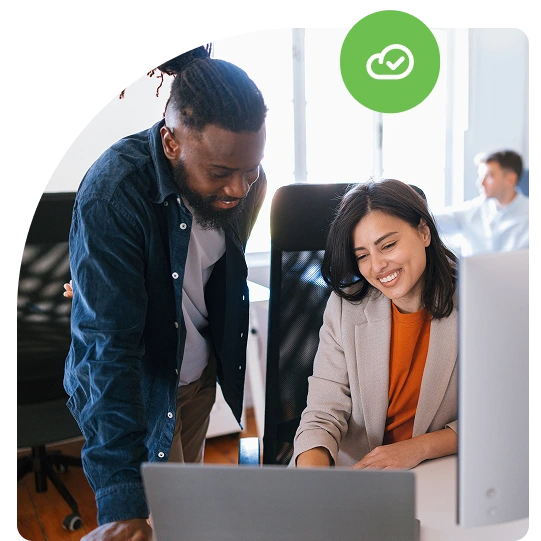 Two coworkers smile at a laptop screen, with a green checkmark-cloud icon overlaid in the corner