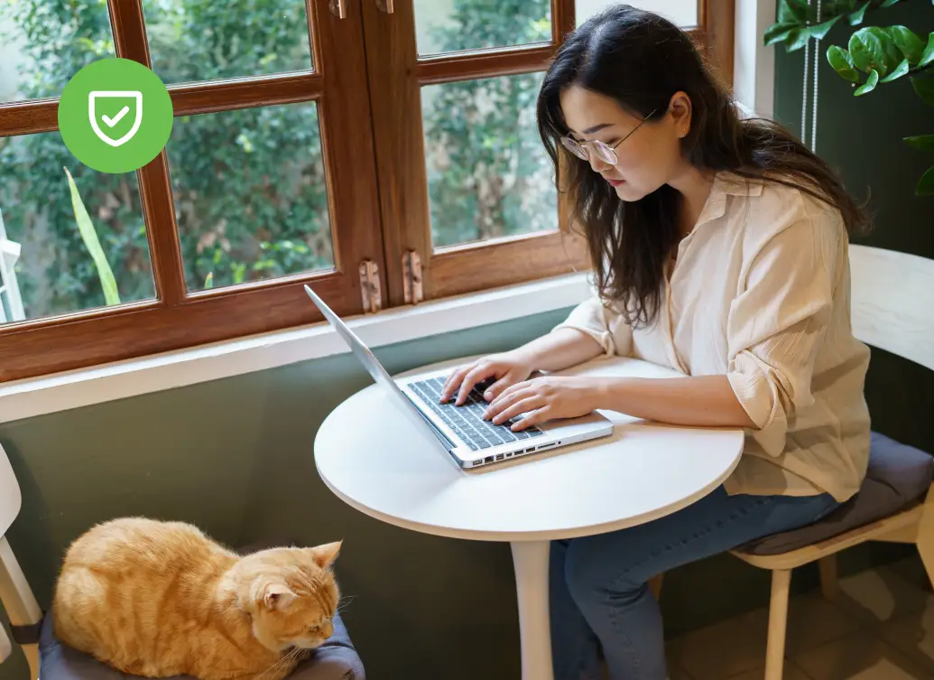 Woman types on a laptop at a small table by a window with a cat sitting nearby