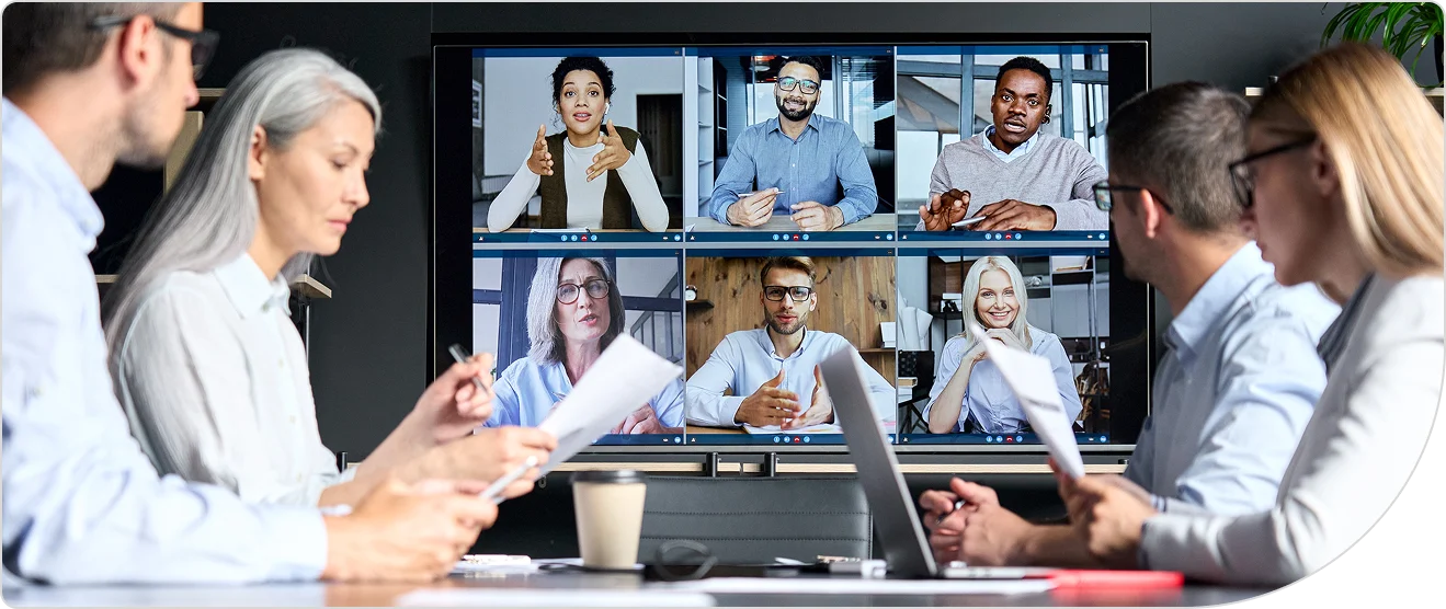 A group of work colleagues sitting around a boardroom tables with laptops and documents and a video conference call screen in the background with 6 people on the video screen or monitor
