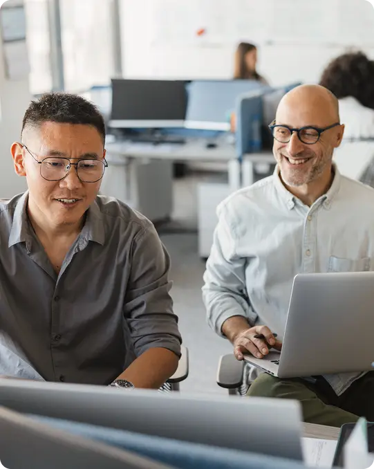 Two colleagues smiling while working at their computers in a bright office, with other employees focused at desks in the background