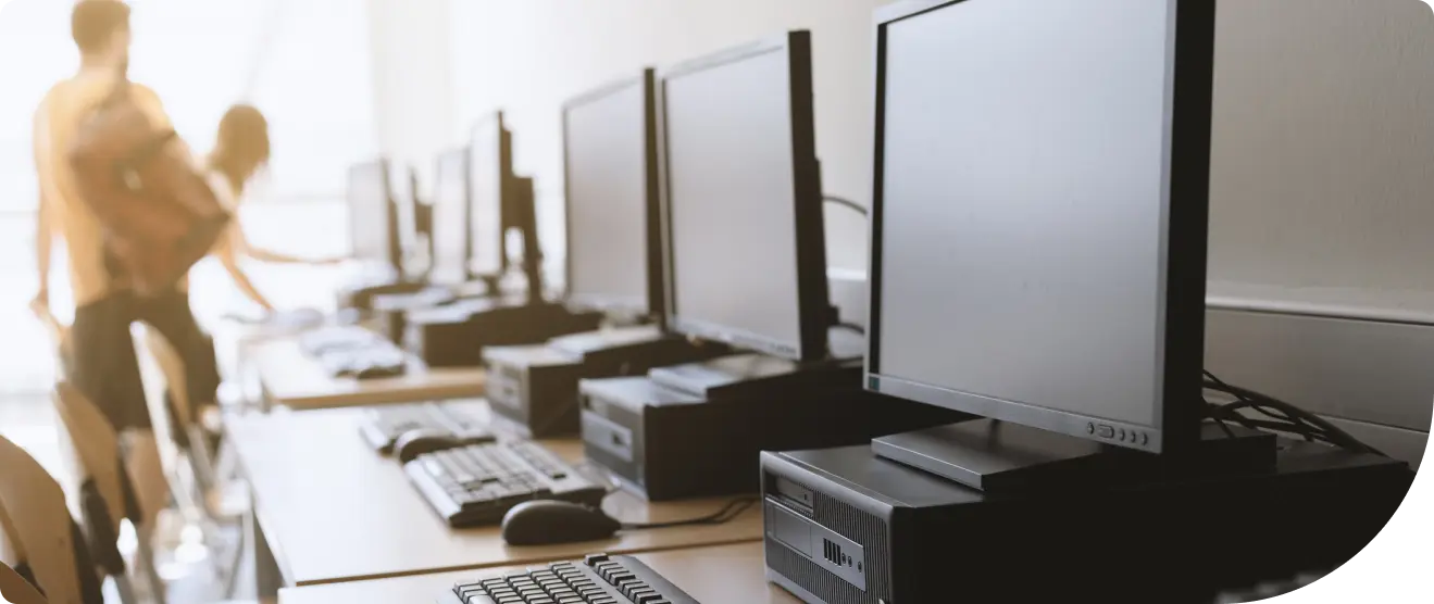 A row of desktop computers with monitors, keyboards, and mice set up on desks in a computer lab. In the background, two people stand near windows