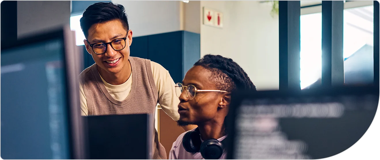 Two men looking at a computer monitor, one standing beside the other one that is sitting