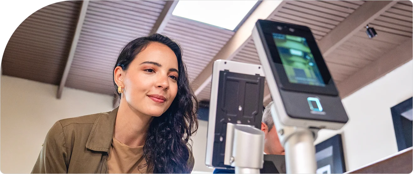 A woman smiling at a biometric access control kiosk with facial recognition scanner in a modern office lobby