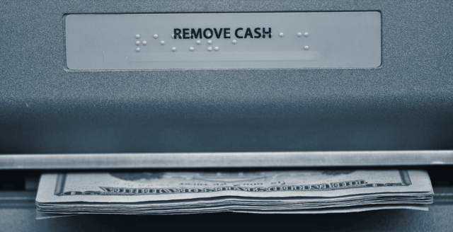 An extreme closeup of an ATM shows a label that says Remove Cash and a stack of twenties emerging.